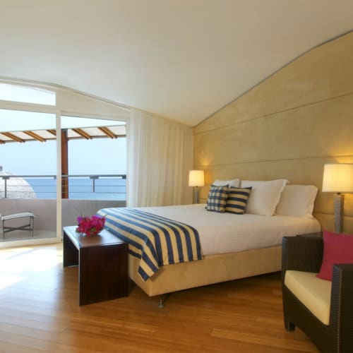 A modern hotel room with a large bed, sitting area, and balcony overlooking the ocean.