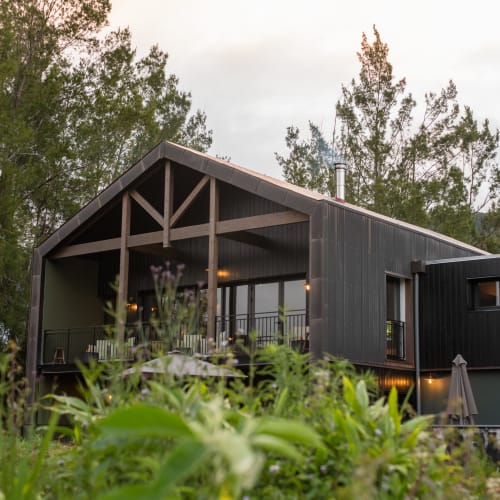 Schwarzes modernes Holzhaus mit großem Balkon, umgeben von üppigen grünen Bäumen und Pflanzen unter einem bewölkten Himmel.