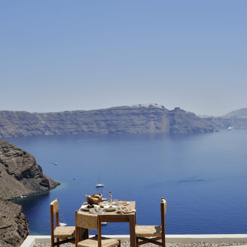 A wooden table with food and drinks set for two on a terrace overlooking a blue sea and rocky cliffs under a clear sky.