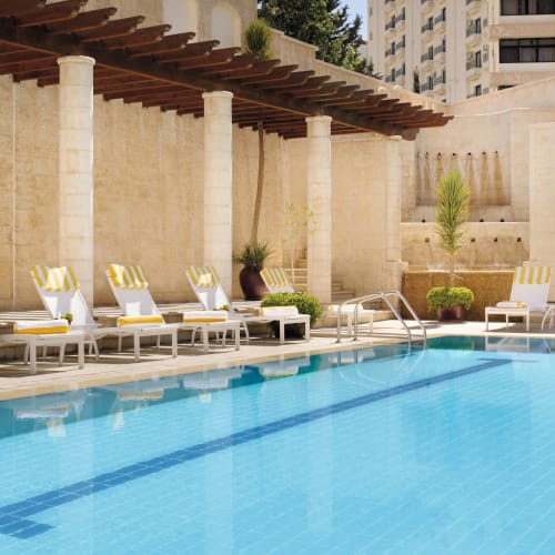 Ein luxuriöser Poolbereich im Moevenpick Resort Petra mit Liegestühlen mit gelb-weiß gestreiften Kissen, umgeben von beige Steingemäuern und einer Holzpergola.