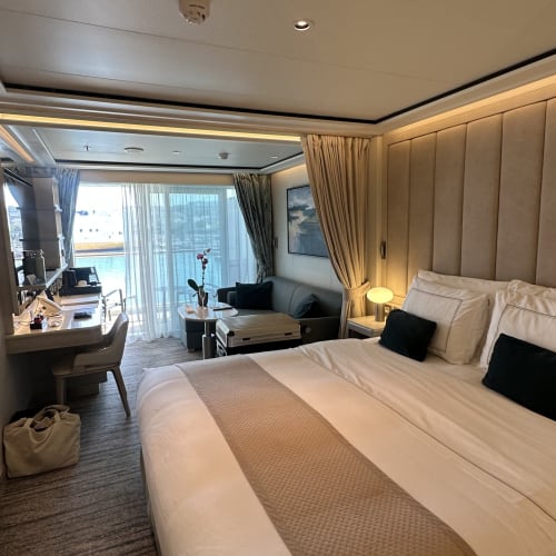 Luxuriöse Kabinensuite auf einem Kreuzfahrtschiff mit großem Bett, Schreibtisch, kleiner Sitzgruppe mit Sofa und Couchtisch sowie Balkon mit Meerblick.