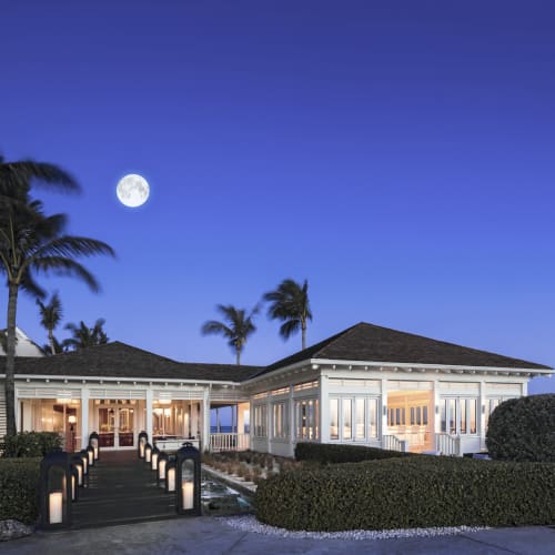 Ein luxuriöses Restaurant am Meer mit großen Glasfenstern, Palmen und Vollmond am Nachthimmel.