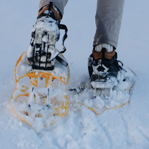 Close-up of a person's feet wearing snowshoes while walking on snow, with snow clinging to the shoes and legs in beige pants.