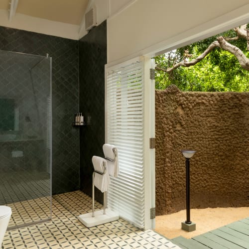 Modernes Badezimmer mit Duschabtrennung aus Glas, weißer Toilette und gemusterten Bodenfliesen, das zu einem Außenbereich mit einem Lehmwand und Grünflächen führt.