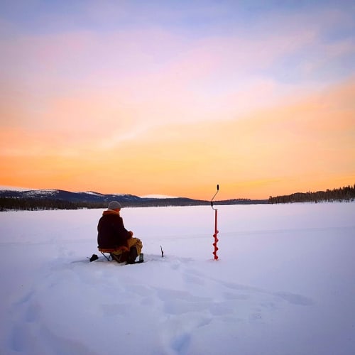 Eine Person sitzt auf einem kleinen Hocker und angelt im Eis auf einem zugefrorenen See während eines farbenfrohen Sonnenuntergangs in einer verschneiten, bewaldeten Landschaft in Finnland.