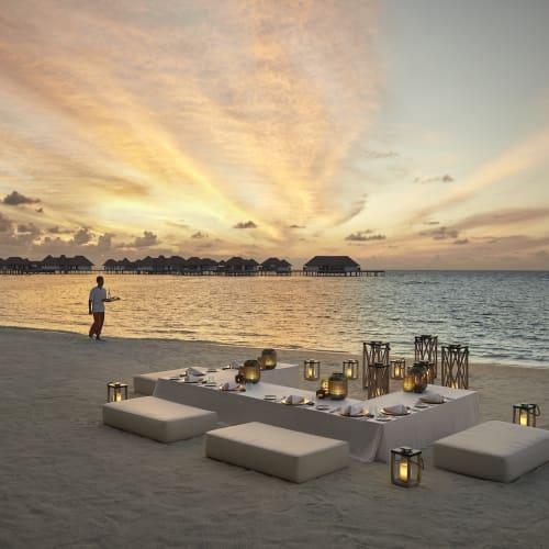 A beachside setup with low tables and cushions for dining during sunset, with a person walking along the shore and overwater bungalows in the background.