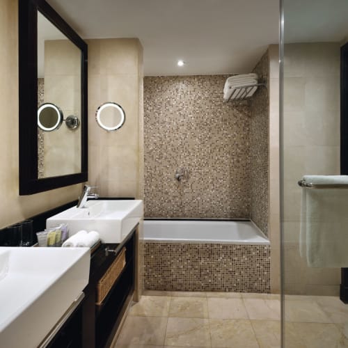 Modern bathroom with two sinks, a large mirror, a bathtub with a mosaic tile surround, and a frosted glass door.