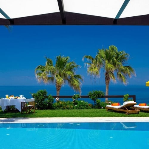 Eine luxuriöse Außenterrasse mit Pool, Blick auf das Meer, mit Liegen, einem gelben Sonnenschirm, einem Esstisch und Palmen in Griechenland.