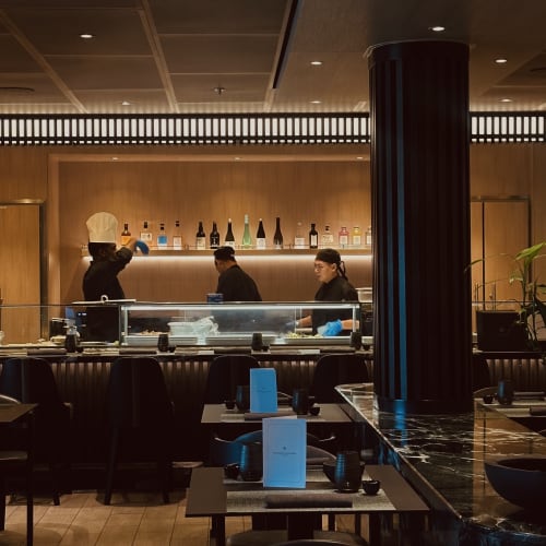 Koch und Küchenpersonal arbeiten hinter einer Theke in einem modernen, gedämpft beleuchteten Restaurant mit Flaschen auf einem hölzernen Wandregal und einer Marmor-Theke im Vordergrund.