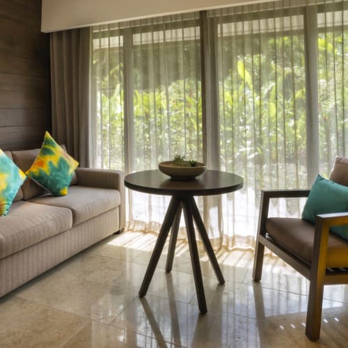 A cozy living room with a beige sofa decorated with colorful cushions and a wooden armchair, set against large windows with sheer curtains showing greenery outside.