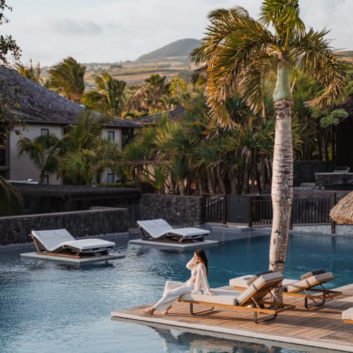 Eine Frau in weiß sitzt auf einem Liegestuhl neben einem Pool, umgeben von Palmen und tropischer Vegetation in einem Resort.