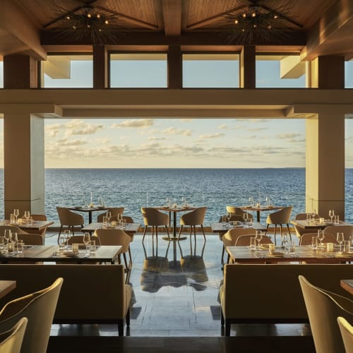 Elegantes Restaurant-Innenraum mit Meerblick durch große offene Fenster und ordentlich gedeckte Tische und Stühle.