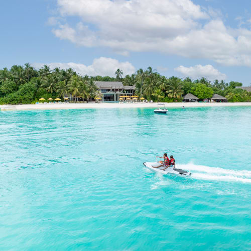 Zwei Personen fahren mit einem Jetski auf klarem türkisfarbenem Wasser nahe einer tropischen Insel mit Palmen und Sonnenschirmen.