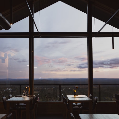 Interior of a modern restaurant with large floor-to-ceiling windows offering a panoramic view of a landscape under a cloudy sunset sky.