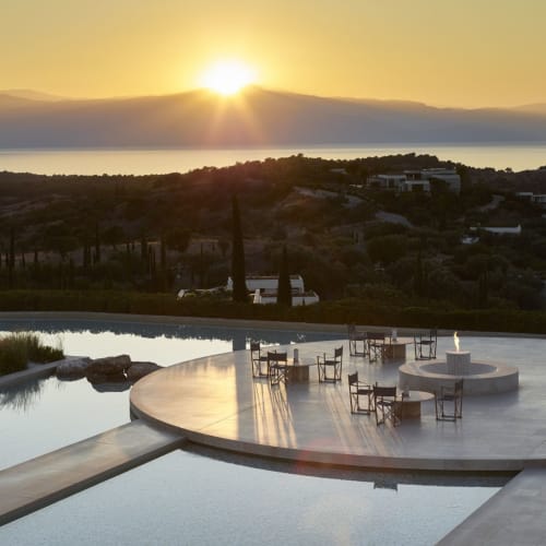 Ein Panorama blick auf eine luxuriöse Outdoor-Lounge-Terrasse bei Sonnenuntergang, mit Blick auf sanfte Hügel, vereinzelt stehende Bäume und moderne Gebäude im Hintergrund.