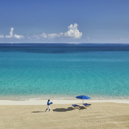 A person walking on a sandy beach towards the calm turquoise ocean with a blue umbrella and two lounge chairs nearby.