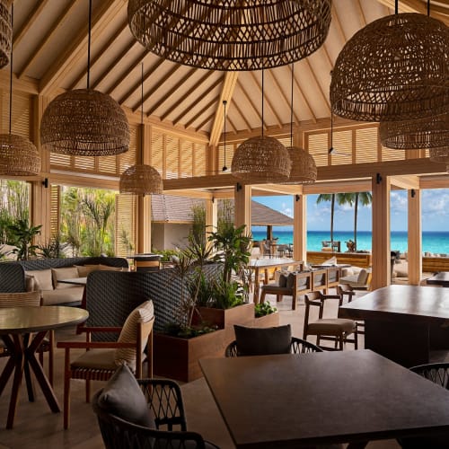 Inside a tropical restaurant with large hanging wicker lamps, overlooking a beach with umbrellas and palm trees.
