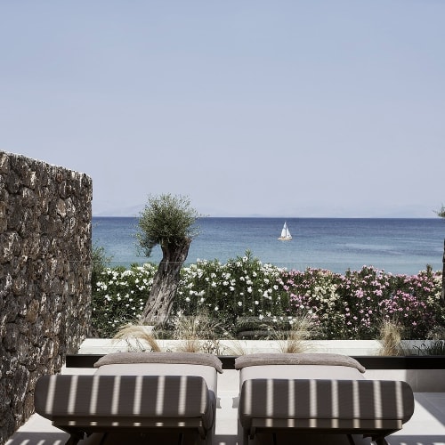 Two lounge chairs on a tiled terrace shaded by a pergola, overlooking a garden with olive trees and flowers, with a sailboat on the sea in the distance under a clear sky.