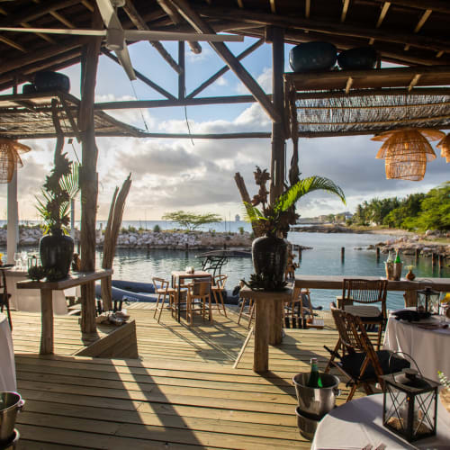 A waterfront restaurant terrace with elegantly set tables and tropical decorations, overlooking the sea and lush greenery.