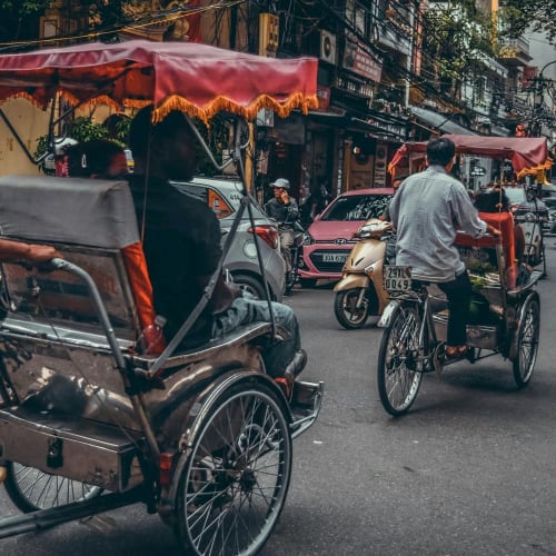 Rikschafahrer und Fahrradfahrer befahren eine belebte Straße in Hanoi, Vietnam, mit Geschäften und Motorrädern entlang der Straße.