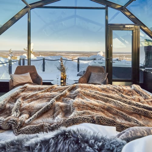 Luxuriöses Schlafzimmer mit Pelzdecken, zwei Sesseln und großen Glasfenstern mit Panoramablick auf eine verschneite Landschaft.