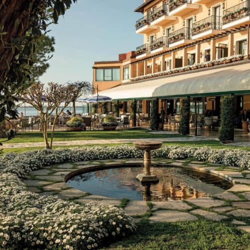 Ein malerischer Blick auf einen Hotelgarten mit einem kleinen Brunnen, umgeben von weißen Blumen und ordentlich geschnittenen Büschen, mit einem mehrstöckigen Hotelgebäude im Hintergrund.