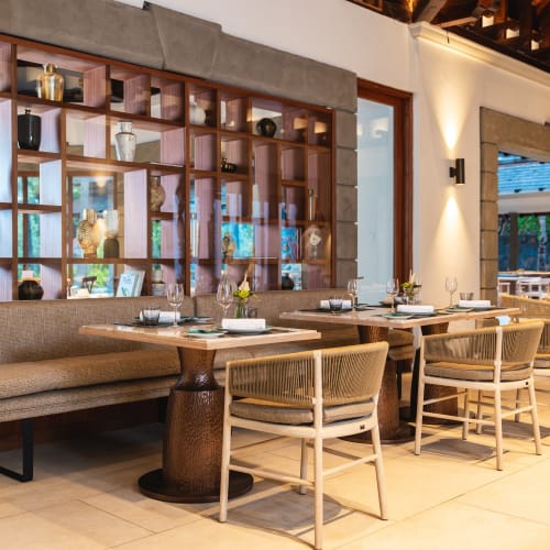 Essbereich in einem Resort-Restaurant mit Holztischen, beige gepolsterter Bank und Stühlen, dekorativen Regalen mit Vasen und Ornamenten sowie sanfter Beleuchtung.