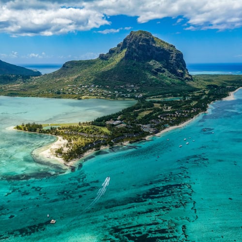 Aerial view of a lush tropical island with a prominent mountain peak, surrounded by turquoise waters and coral reefs under a partly cloudy sky.