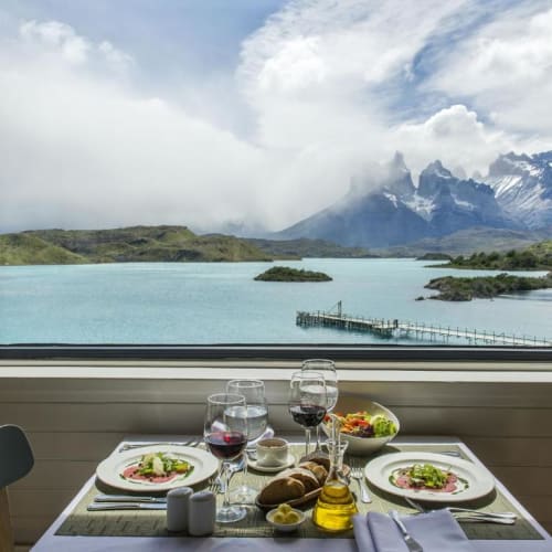 A beautifully set dinner table by a large window overlooking a scenic lake and mountains in Patagonia.