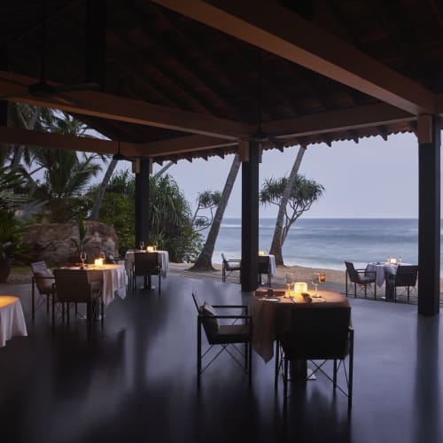 Ein ruhiger Strandrestaurant mit Tischen und Stühlen unter einem Dach, mit Blick auf den Ozean, Palmen und Wellen.