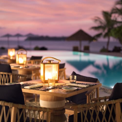 Outdoor dining area at sunset with lit lanterns, tables set with utensils, overlooking a swimming pool and tropical landscape.