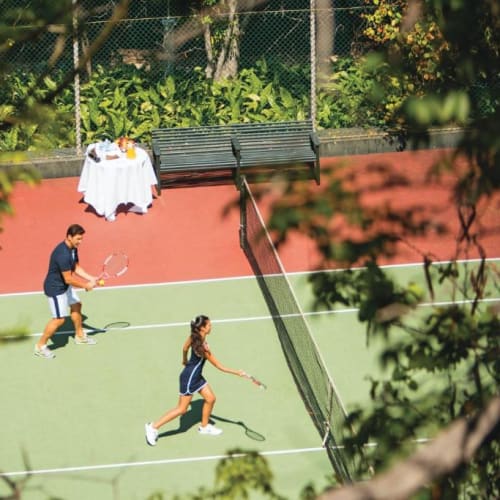 Two children playing tennis on an outdoor court surrounded by greenery and a pink wall.