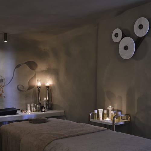 Cozy spa room with a massage table covered in a brown towel, dim candlelight on a counter, and spa products on a trolley against beige walls with soft lighting.