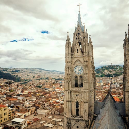 A tall gothic-style church clock tower rises above the city of Quito, Ecuador, with a cloudy sky overhead.