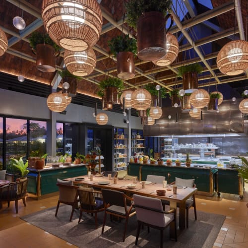 Innenbereich eines Restaurants mit großen Fenstern und Sonnenuntergangsblick, mit Holztischen und -stühlen, hängenden Korblampen und von der Decke hängenden Topfpflanzen.