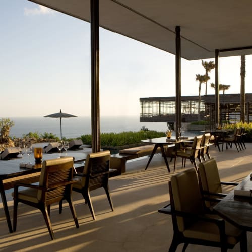 Upscale restaurant with outdoor seating overlooking the ocean, with tables set for dining and surrounded by palm trees.