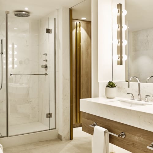 Modern hotel bathroom with a glass shower enclosure, a double vanity with a large mirror, and a view of a balcony outside.