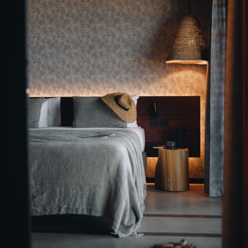 Gemütliches Schlafzimmer mit einem ordentlich gemachten Bett, bedeckt mit einer grauen Decke, einem Strohhut auf gestreiften Kissen, einem hölzernen Nachttisch mit Kessel und Tasse sowie einer gewebten Lampe an einer strukturierten beigen Wand.