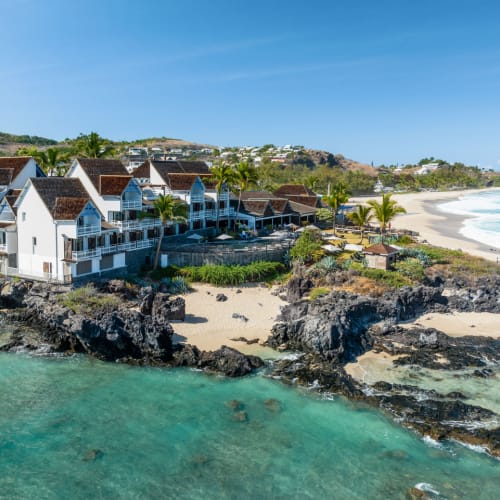 Küstenresort-Gebäude mit roten Dächern auf felsigen Klippen neben einem türkisfarbenen Meer und einem Sandstrand unter klarem blauem Himmel.