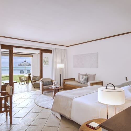 Bright, airy beachfront hotel room with a large bed, wooden furniture, and sliding glass doors opening to a patio with table, chairs, and umbrella overlooking the ocean.