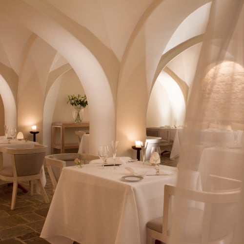 Elegant restaurant interior with white arches, draped curtains, and neatly set tables with white tablecloths and candles.
