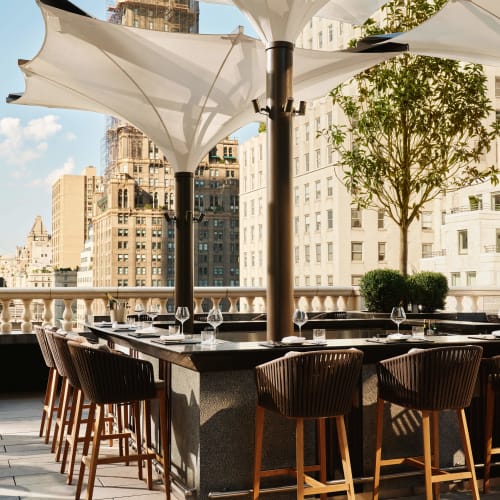 Outdoor dining area on a rooftop terrace in New York City with large white umbrellas, tall buildings, and a tree.