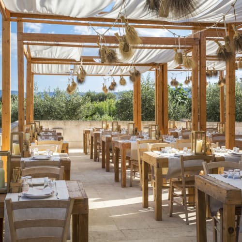 A spacious outdoor restaurant terrace with wooden tables and chairs, decorated with hanging dried herbs and candles, overlooking a lush garden.