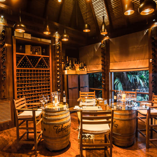Cozy wine bar with wooden furniture, wine bottles, glasses, and barrel tables, illuminated by warm lighting, overlooking a tropical view.