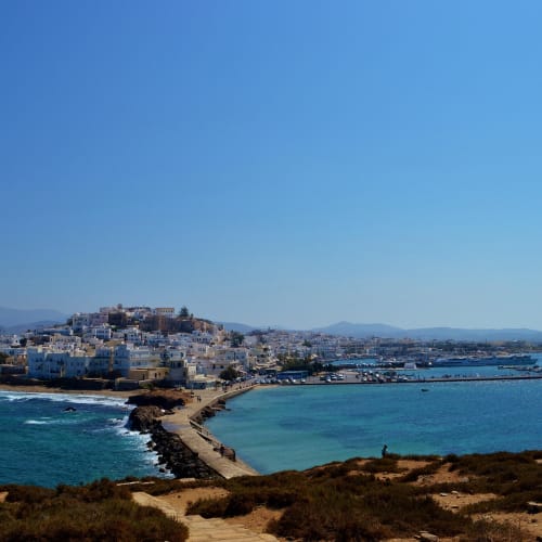 Coastal view of Naxos town with white buildings on a hill, surrounded by blue sea and clear sky, connected by a narrow pier to the mainland with hills in the background.
