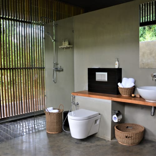A modern bathroom with an open shower area, a wall-mounted toilet, and a wooden countertop with a vessel sink, surrounded by wicker baskets and a large window revealing greenery outside.
