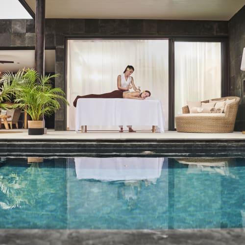 Eine Frau erhält eine Massage auf einem Tisch am Pool eines modernen Lodges mit dunklen Steinwänden, Korbmöbeln und Topfpflanzen.