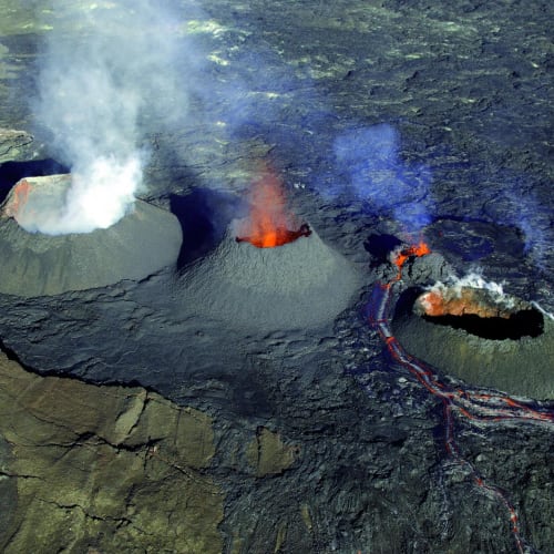 Luftaufnahme von drei Vulkankegeln mit Fumarolen und Lavaflüssen, die bei einem aktiven Vulkanausbruch auf einer kargen Landschaft austreten.