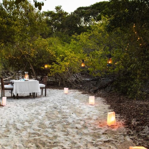 Ein romantisches Privatabendessen auf einem sandigen Strand, umgeben von üppigem Grün mit Kerzen in Laternen und Kerzenhaltern.
