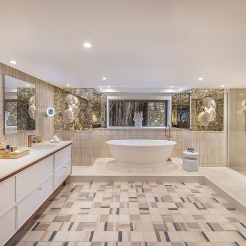 Spacious modern bathroom with a freestanding bathtub centered under a window, a large vanity with double sinks and mirrors on the left, and a glass-enclosed shower on the right, featuring warm ambient lighting and decorative touches.